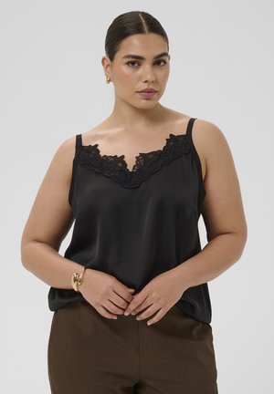 Femme aux cheveux foncés coiffés en chignon lisse, portant un caraco noir à bordure en dentelle, un pantalon marron, des boucles d'oreilles créoles dorées et un bracelet en or, les mains jointes.