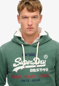 Zielona bluza z kapturem, z białymi i czerwonymi grafikami, z napisem "Vintage SuperDry". Posiada kaptur ze sznurkiem i strukturalny materiał.
