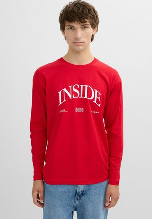 Joven con camiseta roja de manga larga con texto blanco "INSIDE" y vaqueros azul claro, de pie frente a un fondo claro y liso.