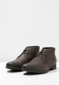 Bottines en cuir marron avec lacets, bouts ronds et talons bas, placées côte à côte sur une surface blanche.