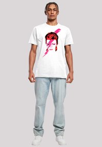 Weißes Baumwoll-T-Shirt mit einem Grafikdruck eines Gesichts mit rotem Haar und einem pinken Blitzdesign vor einem schlichten Hintergrund.
