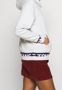 Sudadera gris con capucha y un bolsillo tipo canguro, con un dobladillo de marca en azul marino y rosa. Combinada con pantalones cortos de pana rojos.