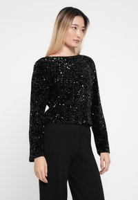 Top negru cu paiete, cu mâneci lungi și decolteu rotund. Suprafață texturată creată de paietele aranjate îndeaproape. Asortat cu pantaloni negri.
