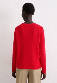 Rode gebreide trui met een ribbelstructuur, ronde halslijn en valt over de schouders, gecombineerd met beige broek, gezien vanuit de achterkant.