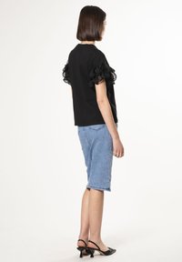 Top negro de manga corta con detalles de volantes en capas, combinado con shorts de denim claros. La modelo lleva sandalias de tacón negras.