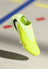 Ciorap de fotbal Nike de culoare galben aprins, cu logo-uri Swoosh negre și roșii, șireturi și talpă cu crampoane, pe un teren de sport interior.