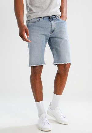 Mand iført lyseblå denimshorts med frynser i kanten, hvide sokker, hvide sneakers og en lys grå t-shirt, står med den ene hånd i lommen.