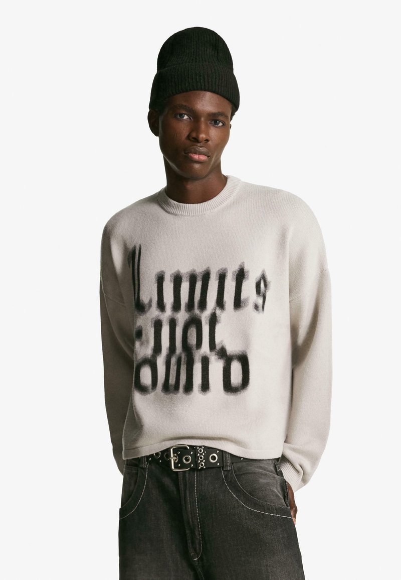 Svetlo šedý pletený svetor s čiernym abstraktným textovým dizajnom, okrúhlym výstrihom a voľným strihom. Vhodný s tmavými džínsami a čiernou rebrovanou čiapkou.