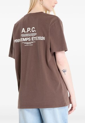 Donna con lunghi capelli biondi che indossa una maglietta marrone A.P.C. Printemps Été 2026 e jeans azzurri, mostrando un tatuaggio sulla schiena e sul braccio.