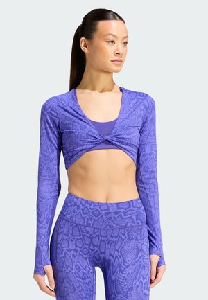 Kvinde iført lilla crop top med slangeprint og leggings med lange ærmer, står med afslappede hænder og ser til højre med neutralt udtryk.