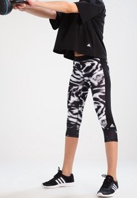 Zwarte cropped sportshirt met korte mouwen; aansluitende leggings met een zwart-wit abstract patroon; zwarte sportschoenen.