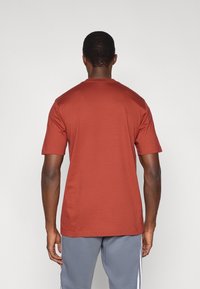 Κοντομάνικο κόκκινο-καφέ βαμβακερό μπλουζάκι με στρογγυλή λαιμόκοψη και ίσια τελική ραφή, φορεμένο με γκρι παντελόνι. Εμφανίζεται η πίσω όψη.