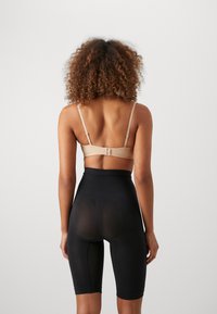 Beige bra with adjustable straps and hook closure, paired with high-waisted black shorts featuring a translucent panel at the rear.