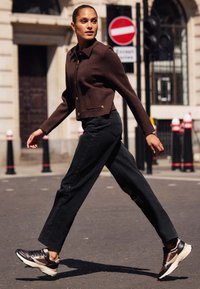 Brązowa krótka kurtka z złotymi guzikami, zestawiona z czarnymi spodniami o wysokim stanie z bocznymi zatrzaskami. Na nogach czarne i beżowe buty sportowe.