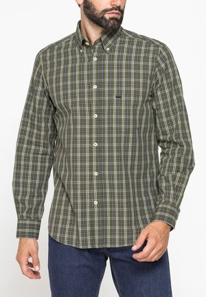 Uomo che indossa una camicia a quadri verde e blu scuro con bottoni e maniche lunghe e jeans blu scuro, rivolto leggermente verso destra.