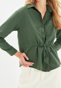 Vrouw in een olijfgroene blouse met knopen en een borstzakje, met een tailleband, gecombineerd met lichtbeige broek, staand met een hand op de heup.