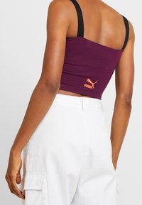 Top corto viola con spalline nere e logo Puma arancione sulla parte bassa della schiena, abbinato a pantaloni cargo bianchi a vita alta.