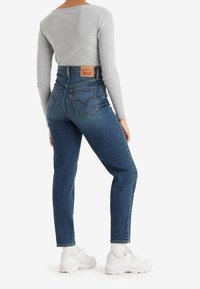 Femme portant un jean taille haute bleu et un haut gris à manches longues, debout de profil, avec des baskets blanches et des chaussettes blanches.