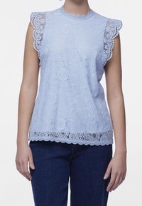 Haut en dentelle bleu clair avec un col et des bords de manches festonnés, présentant des motifs floraux et des détails de tissu texturé. Associé à un jean foncé.