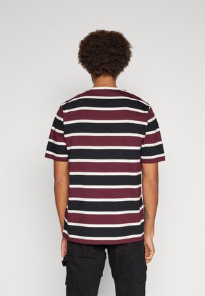 Osoba o kręconych włosach, ubrana w koszulkę z krótkim rękawem w poziome czarno-białe i bordowe paski oraz czarne spodnie cargo, stojąca tyłem.