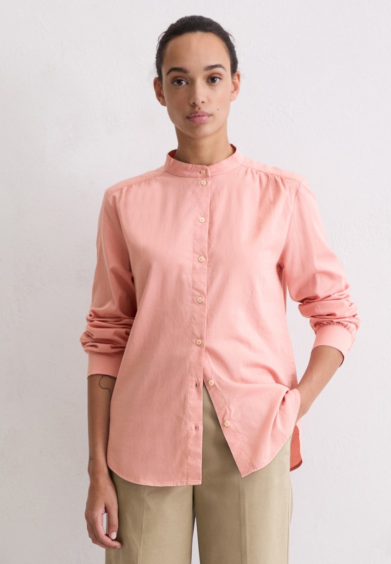 Light pink shirt with a stand-up collar, long sleeves, and button closure. Made from smooth fabric with minimal detailing.