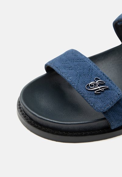 Πέδιλο ανοιχτού τύπου με μαύρη σόλα και ρυθμιζόμενο μπλε υφασμάτινο λουράκι που φέρει μεταλλικό διακοσμητικό λογότυπο σε λευκό φόντο.