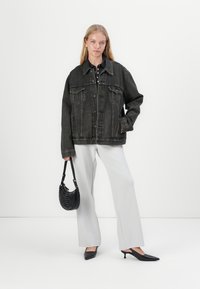 Veste en denim noire oversized avec des fermetures à boutons, deux poches poitrine, associée à un pantalon large blanc et un sac à main noir.