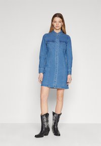 Rochie din denim, albastră, cu deschidere cu nasturi în față, guler, mâneci lungi, două buzunare pe piept, tiv evazat, purtată cu cizme negre în stil western.