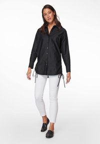 Schwarzes Langarmshirt mit Knopfverschluss, seitlichen Zugbändern und lässigem Schnitt, kombiniert mit weißen Knöchellangen Jeans und schwarzen Slippern.