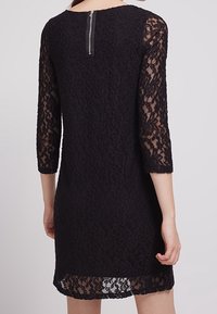 Svart spetsklänning med genomskinliga ärmar som når tre fjärdedelar, en scallopad nederkant och dragkedjestängning i ryggen. Mycket texturerat blommönster.