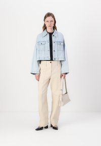 Giacca in denim azzurro corto con due tasche sul petto, indossata su un top nero. Abbinata a pantaloni beige a vita alta e scarpe nere.