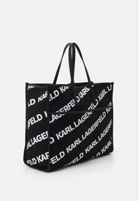 Svart tygväska med vit text "KARL LAGERFELD" i ett upprepat mönster, med två svarta handtag och en strukturerad rektangulär form.