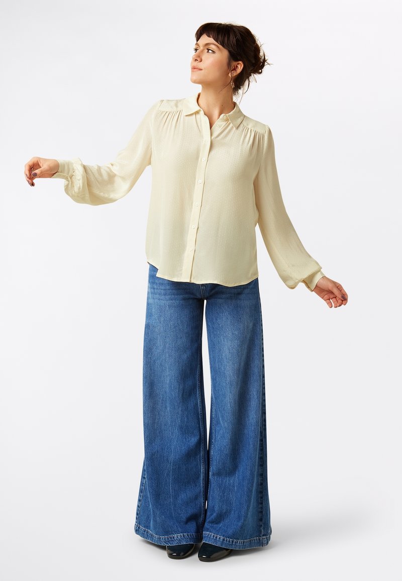 Camicia button-up beige chiaro con maniche a palloncino, abbinata a jeans blu a vita alta e gamba larga in una classica trama di denim.