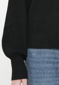 Črn pleteni pulover z rokavom in rebrastim manšetom, ki delno pokriva roko, v kombinaciji s svetloblujimi denim kavbojkami v pasu.