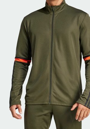 Hombre con chaqueta verde oliva de cremallera completa con rayas negras en las mangas y bandas rojas alrededor de la parte superior de los brazos, combinada con pantalones a juego.