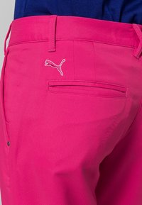 Zbliżenie na kieszeń jasnoróżowych spodni z białym haftem logo Puma, noszone z włożoną do środka ciemnoniebieską koszulą.