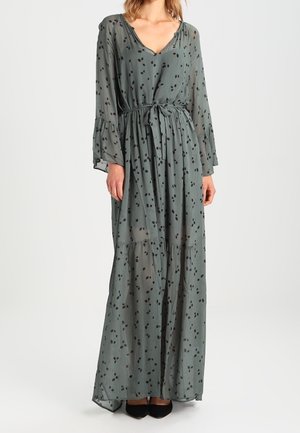 Lang grå kjole med sorte abstrakte pletter, V-hals, lange flagrende ærmer, taljeret med bindebånd, båret med sorte sko.