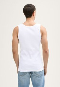 Homme aux cheveux courts et foncés portant un t-shirt sans manches blanc et un jean bleu clair, tourné le dos contre un fond clair.