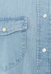 Světle modrá džínsová košile s texturovaným povrchem, náprsní kapsou a stříbrnými patentními knoflíky s perleťovým akcentem.