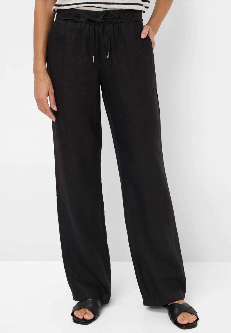 Personne portant un pantalon ample noir à cordon de serrage et des sandales noires à enfiler, debout avec une main dans la poche sur un fond blanc.