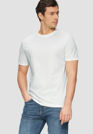 Man wearing plain white short-sleeve t-shirt and blue jeans, standing with one hand in pocket, looking to his left against a plain background.