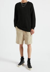 Personne portant un pull noir à manches longues côtelé, un short beige arrivant aux genoux, des baskets montantes noires, des colliers argentés en couches et des bagues à la main droite.