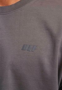 Grå t-shirt med korte ærmer lavet af glat stof, der har et lille prægede "DEF" logo på brystet. Casual design med rund hals.