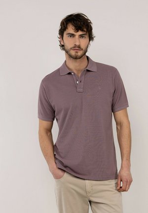 Hombre con cabello corto y rizado que lleva una camisa polo morada apagada y pantalones beige, de pie frente a un fondo liso y claro.