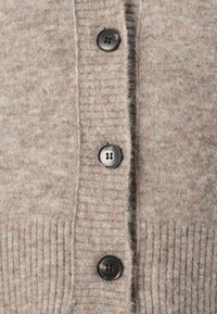 Beige cardigan med en myk tekstur, med fem svarte plastknapper, ribbet kant og et strikkemønster på stoffet.