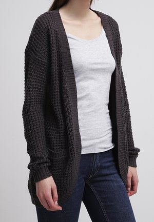 Osoba nosząca ciemnoszary teksturowany dzianinowy kardigan na jasnoszarym dopasowanym t-shircie i ciemnoniebieskich dżinsach, stojąca na jednolitym tle.