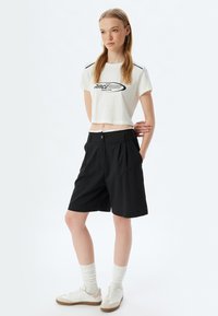 Zwarte op maat gemaakte shorts met plooi, gecombineerd met een witte cropped t-shirt met zwarte tekst en schouderstrepen. Schoenen in lichte kleuren.