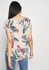 Blouse à manches courtes florale en rose pâle, ornée de feuilles et fleurs tropicales en vert, rouge et blanc. Fermeture zippée dans le dos. Texture lisse.