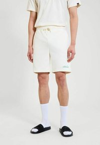 Mitad inferior de una persona que lleva pantalones cortos color crema con cordón, calcetines blancos de canalé y sandalias de goma negras, sobre un fondo liso.