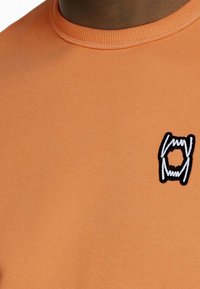 Felpa in cotone arancione con collo a coste e un piccolo logo ricamato in bianco e nero sul lato sinistro del petto.